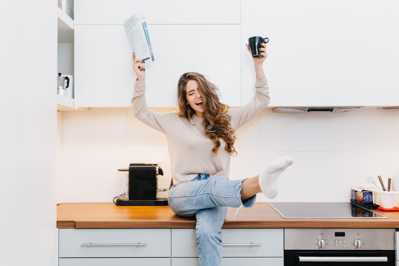 Graceful caucasian girl wears jeans enjoying good morning in her kitchen. Indoor portrait of emotional female model drinking coffee and reading magazine.